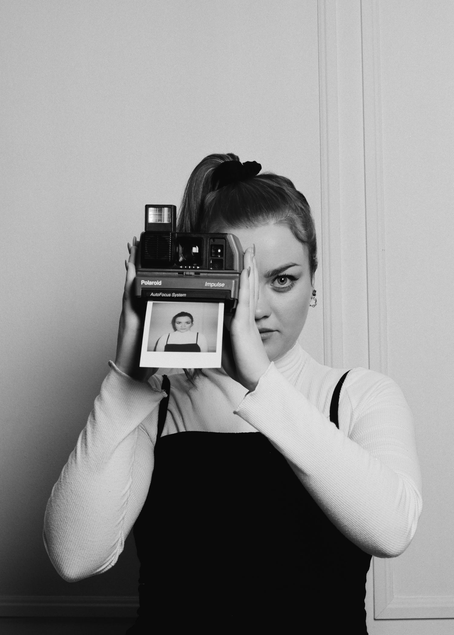 Black and white portrait of photographer Meghan Bond holding a Polaroid camera in front of her face, with a self-portrait emerging from the film — captured by Toronto-based photographer Nicholas Hyatt in a minimal, editorial style.