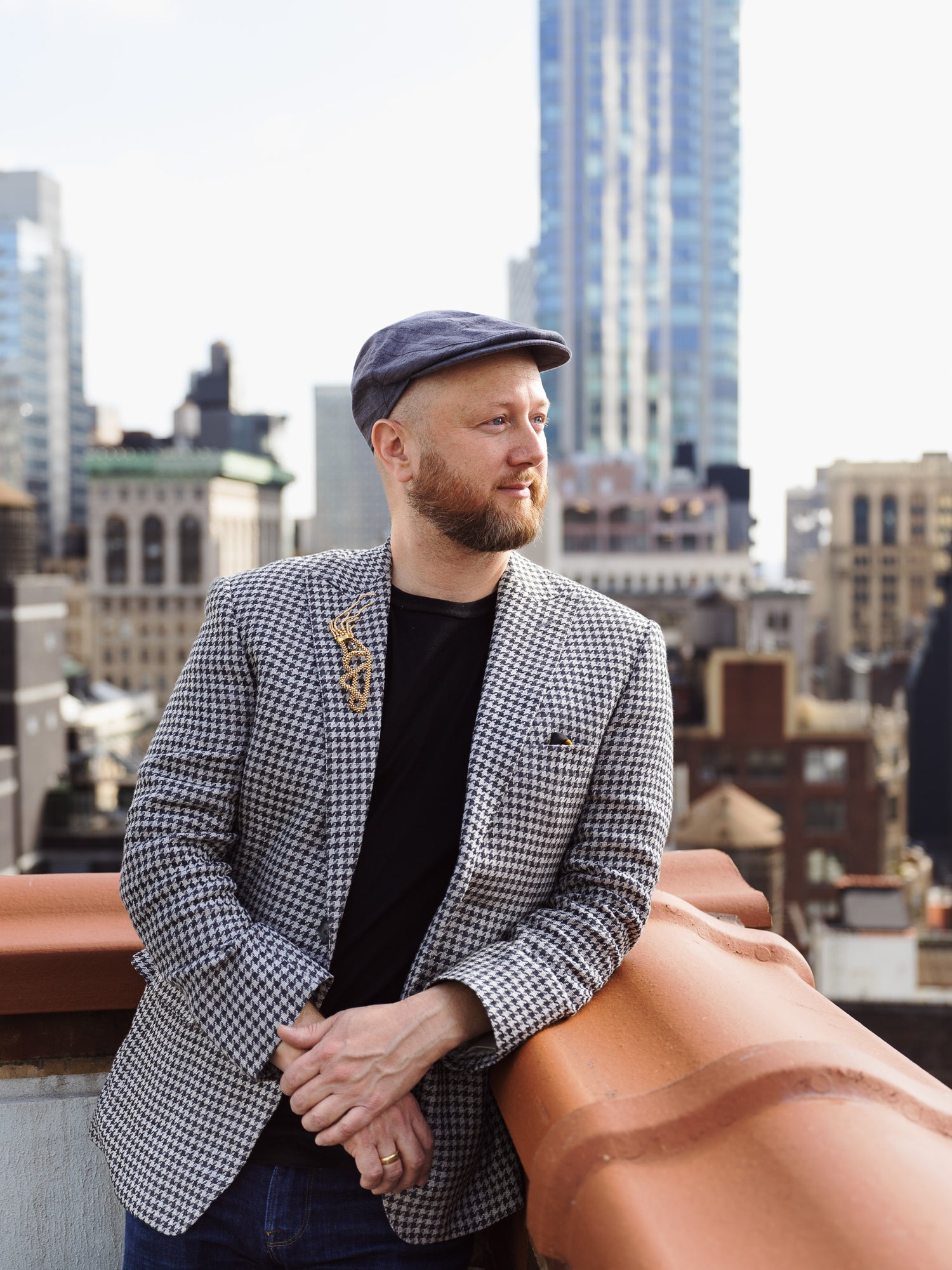 Jeweller Chris Davies wearing a trimmed beard and flat cap leans against a rooftop ledge, wearing a black shirt and houndstooth blazer adorned with a gold brooch, with a city skyline rising in the background. Photographed by Toronto-based photographer Nicholas Hyatt, this editorial portrait captures personality and urban sophistication in natural light.