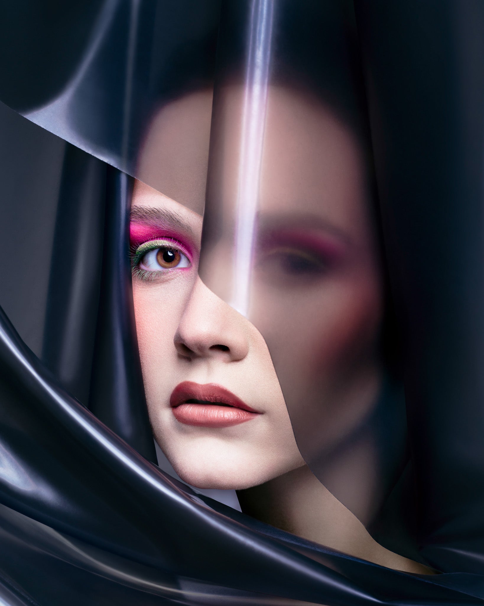 A striking beauty portrait of a woman with bold pink and green eyeshadow and matte red lips, partially veiled in translucent black plastic. Captured by Toronto-based photographer Nicholas Hyatt, the image merges avant-garde styling with modern studio beauty.