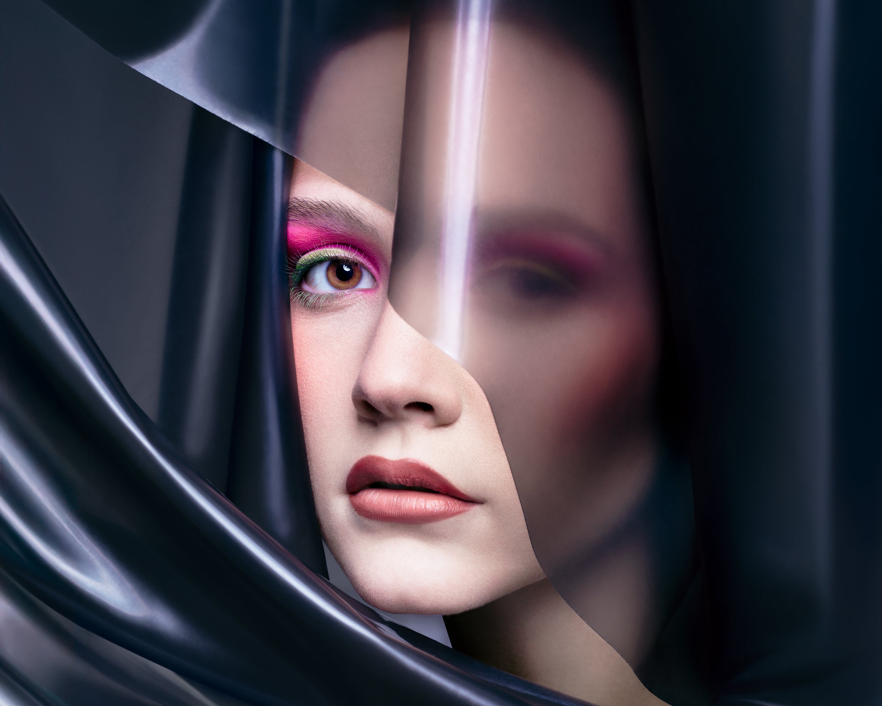  A striking beauty portrait of a woman with bold pink and green eyeshadow and matte red lips, partially veiled in translucent black plastic. Captured by Toronto-based photographer Nicholas Hyatt, the image merges avant-garde styling with modern studio beauty.