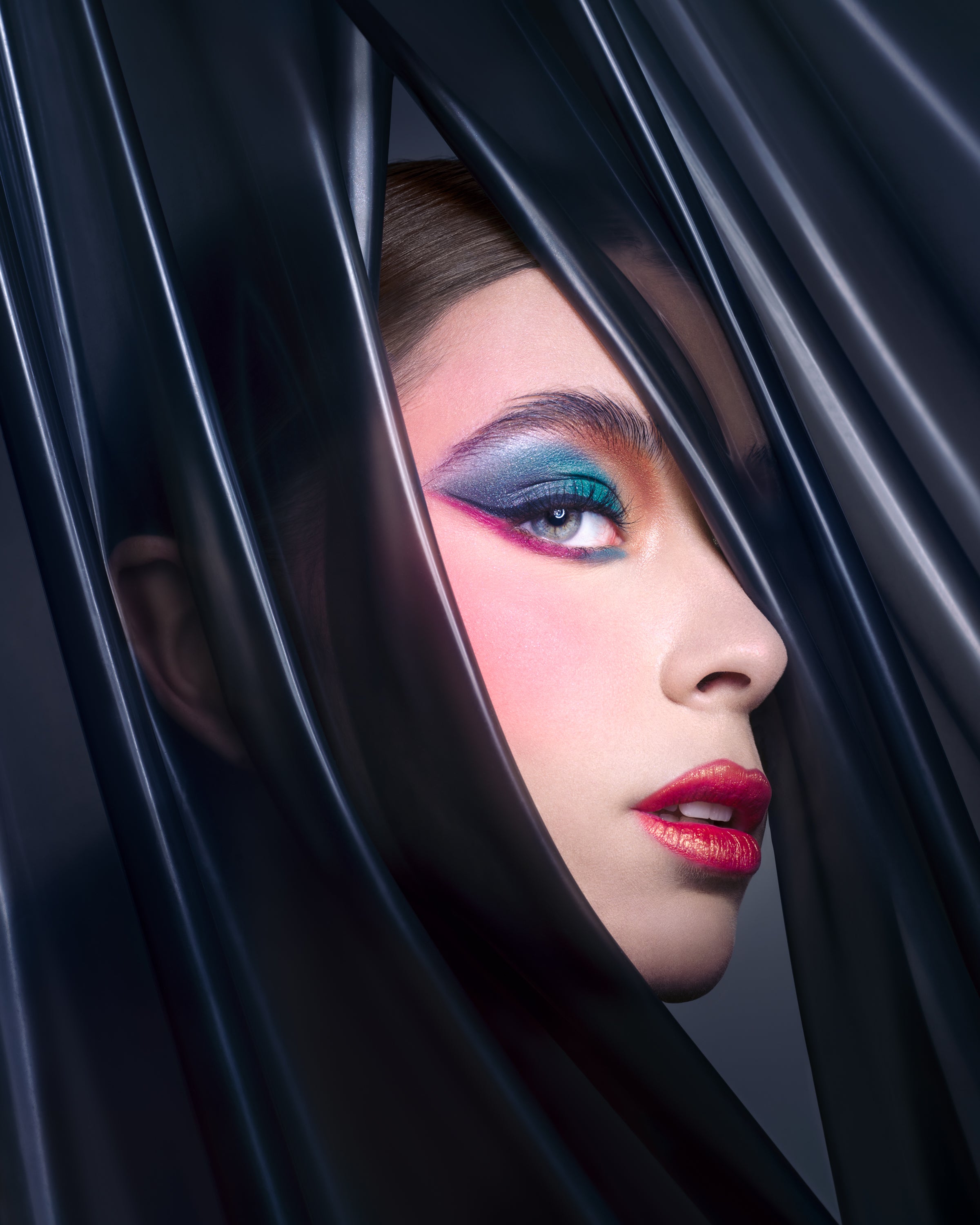  A close-up of a model’s face with bold teal, magenta, and blue eyeshadow and red lips, framed by glossy black latex. Photographed by Toronto-based beauty photographer Nicholas Hyatt, this editorial image celebrates color contrast and high-impact styling.