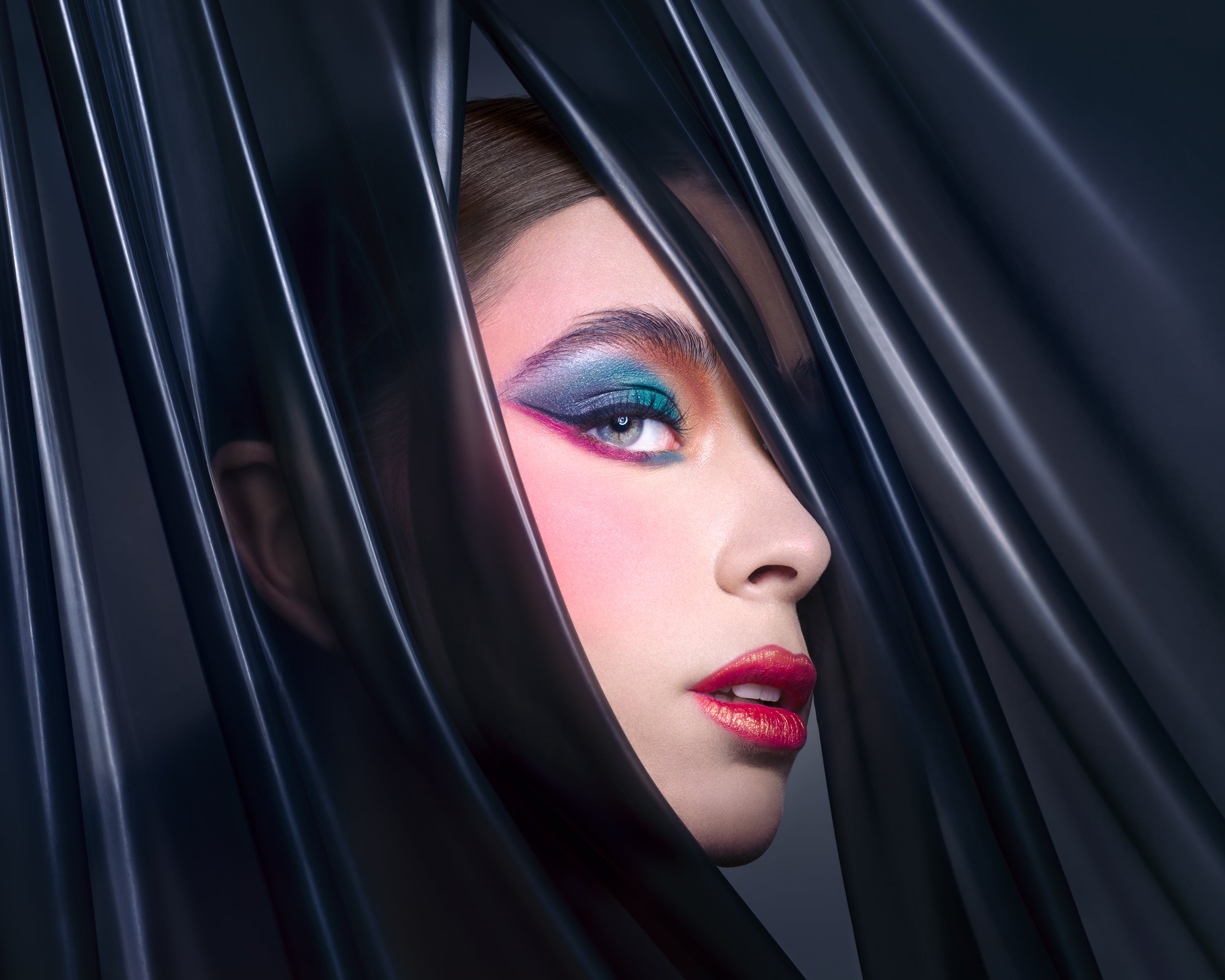  A close-up of a model’s face with bold teal, magenta, and blue eyeshadow and red lips, framed by glossy black latex. Photographed by Toronto-based beauty photographer Nicholas Hyatt, this editorial image celebrates color contrast and high-impact styling.