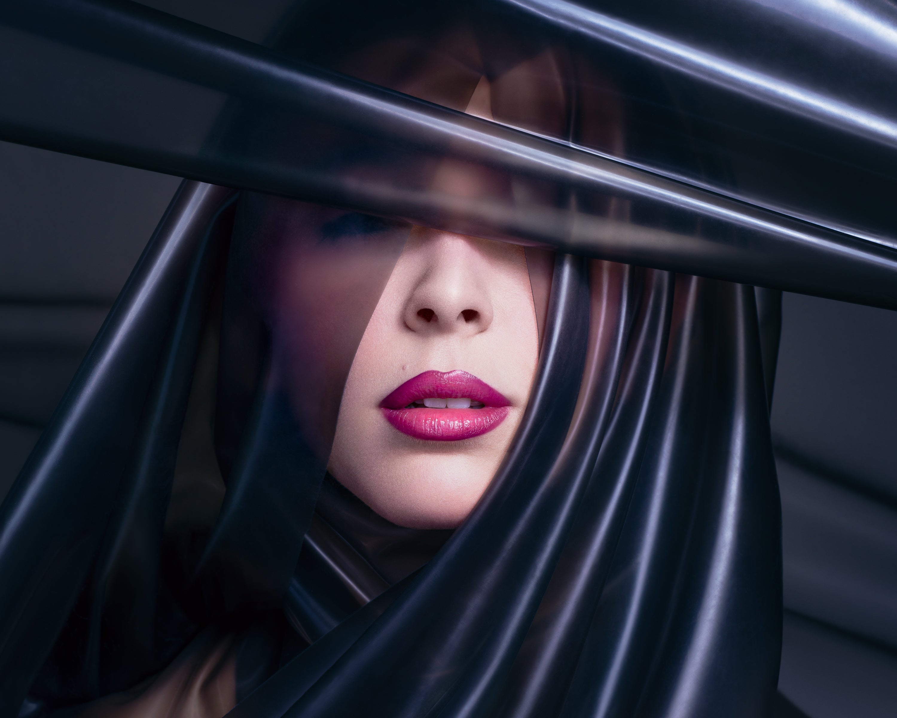 A mysterious close-up beauty portrait of a woman with glossy magenta lips, partially veiled by layers of sheer black plastic. Captured by Toronto-based photographer Nicholas Hyatt, blending sensuality with conceptual beauty photography.
