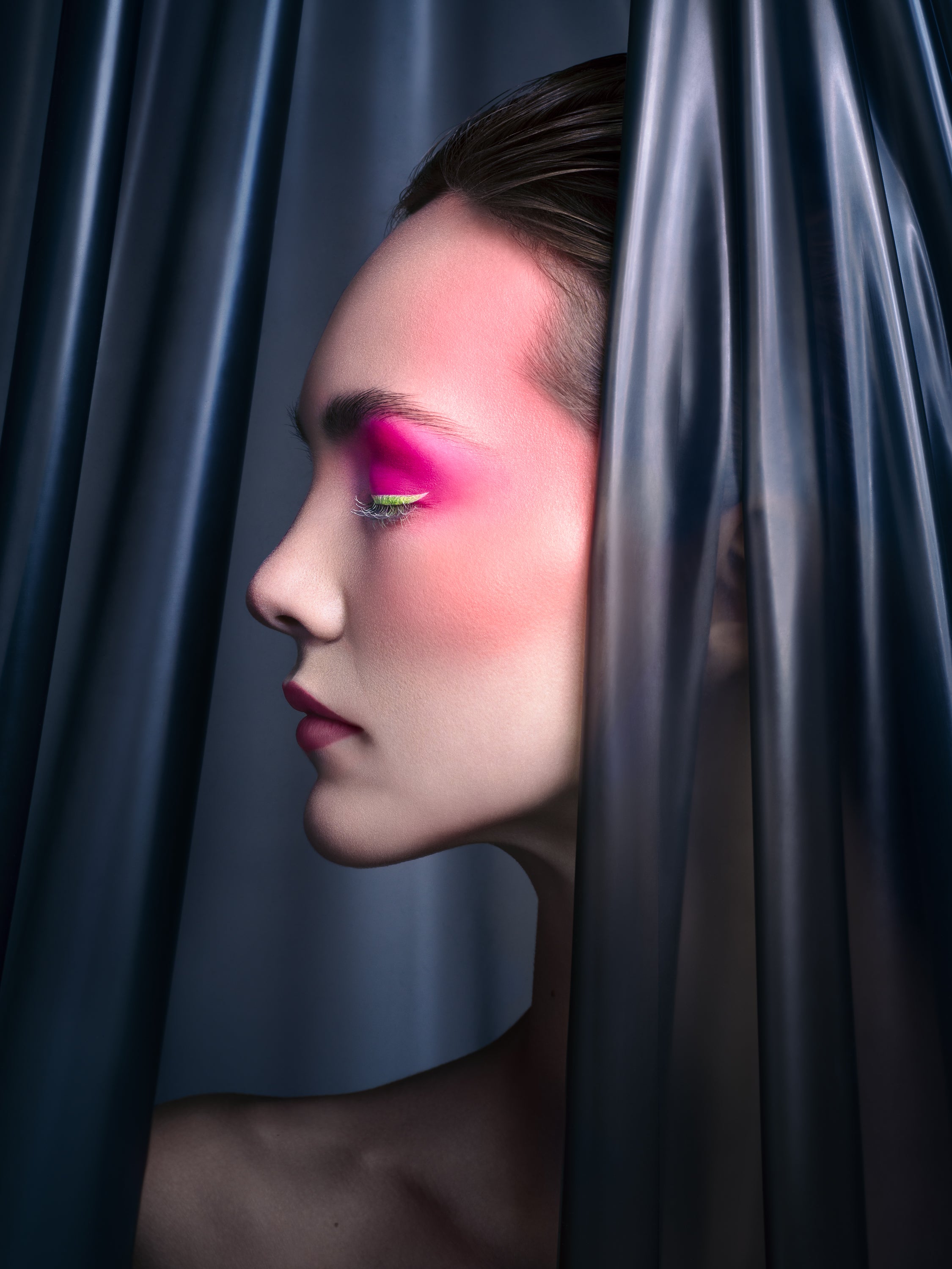 Profile view of a model with neon pink and green eyeshadow, soft matte lips, and flawless skin, framed by dark translucent drapery. Photographed by Toronto-based beauty photographer Nicholas Hyatt, this image emphasizes shape, color, and form in modern beauty styling.