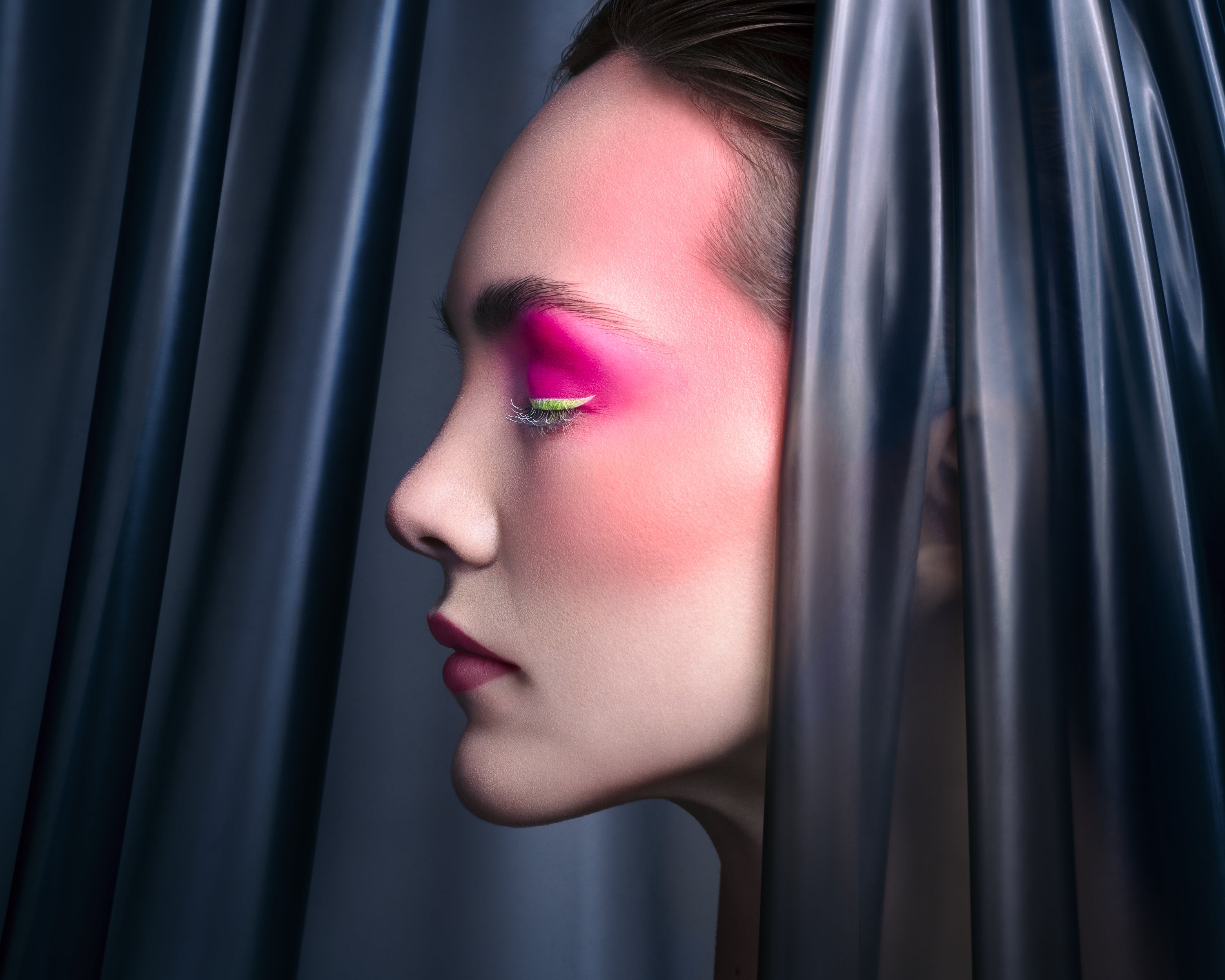 Profile view of a model with neon pink and green eyeshadow, soft matte lips, and flawless skin, framed by dark translucent drapery. Photographed by Toronto-based beauty photographer Nicholas Hyatt, this image emphasizes shape, color, and form in modern beauty styling.