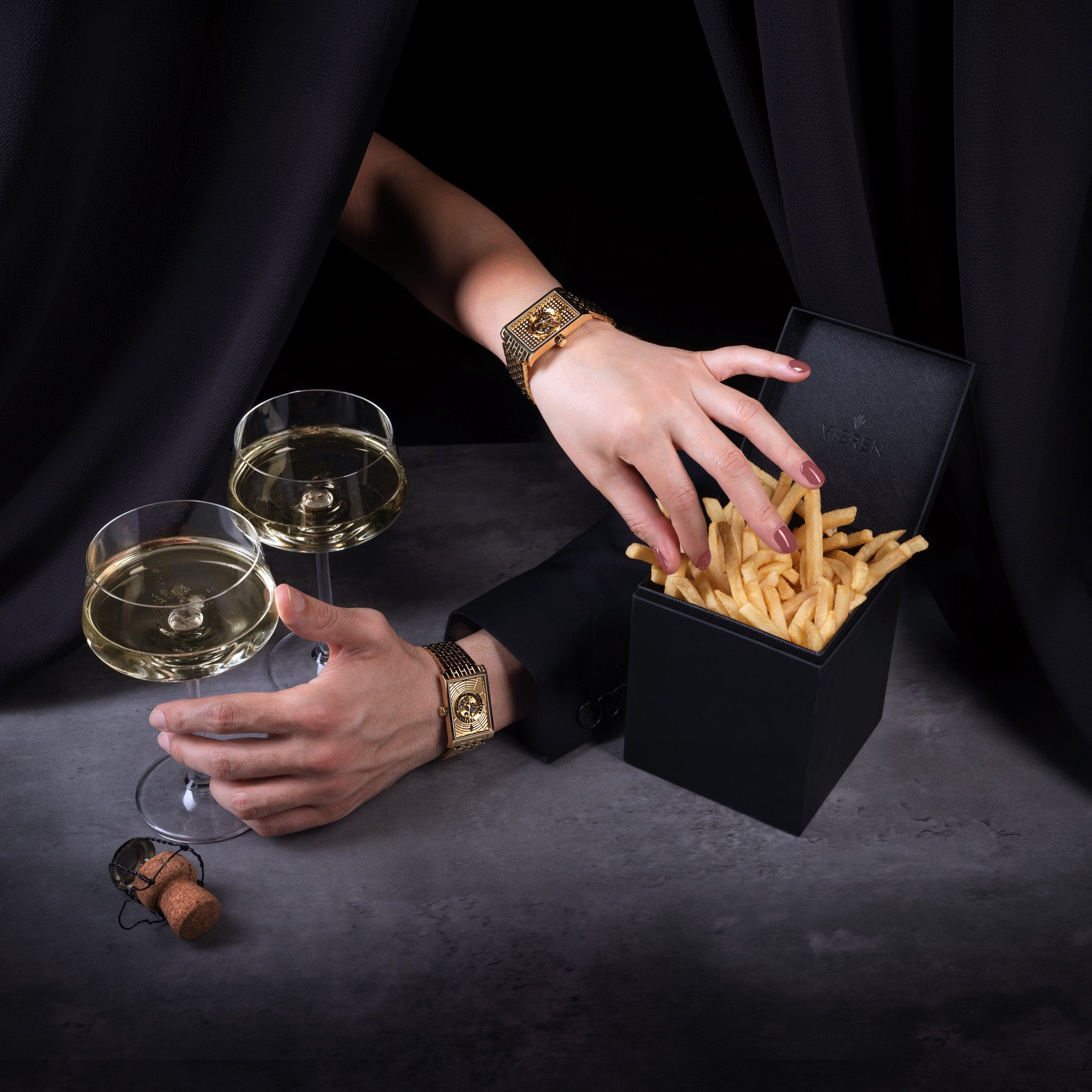 A luxurious still life featuring two hands adorned with gold VIEREN watches reaching for a sleek black box of French fries beside two coupe glasses of champagne. Photographed by Toronto-based photographer Nicholas Hyatt, this image blends indulgence and elegance in a playful, fashion-forward editorial style.
