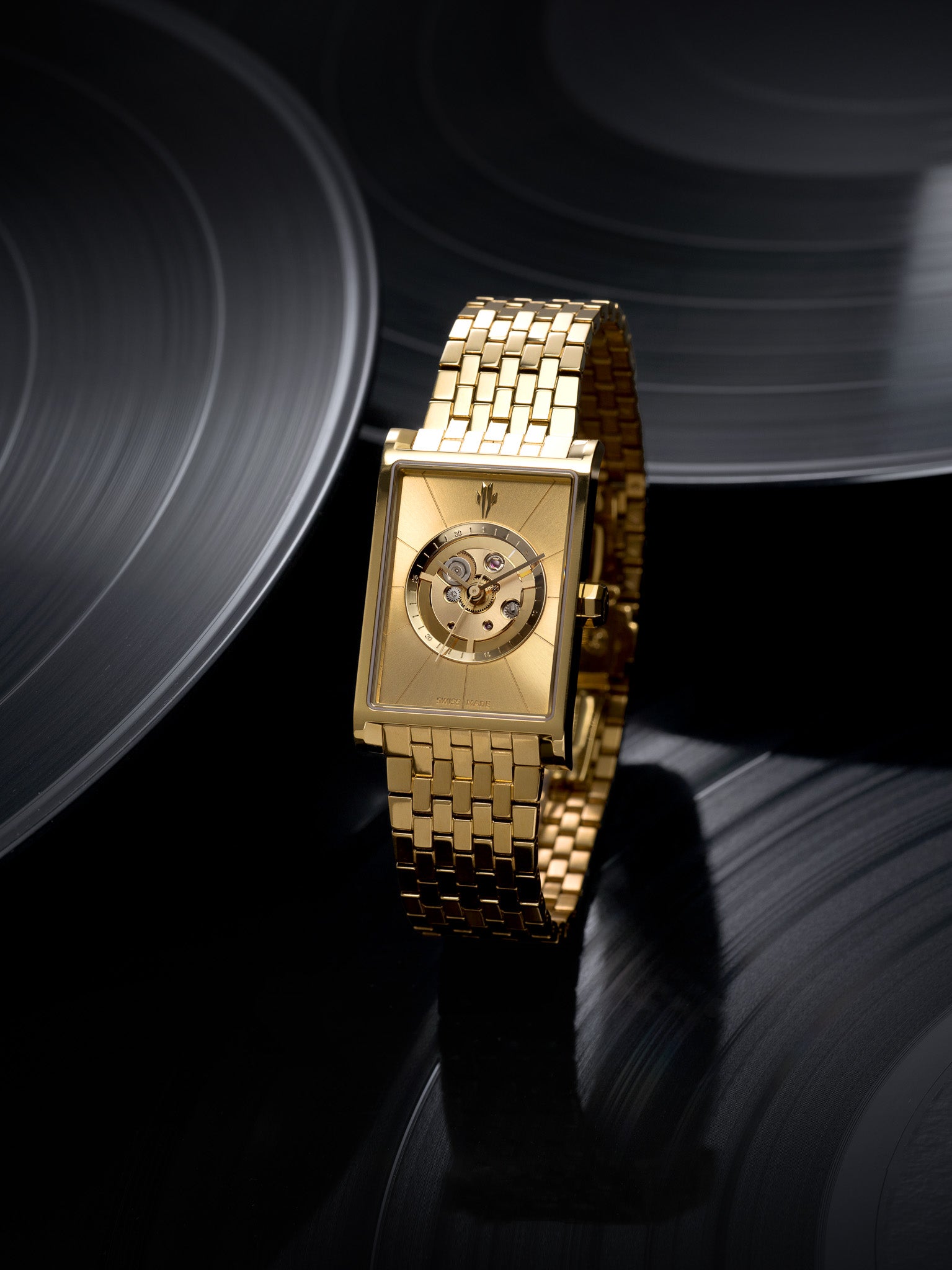 A gold Vieren automatic watch with a rectangular face and exposed movement is showcased on a reflective black surface styled to resemble vinyl records. The Swiss-made timepiece exudes luxury and precision. Photographed by Toronto-based commercial photographer Nicholas Hyatt, this still life highlights high-end craftsmanship with a music-inspired visual theme.