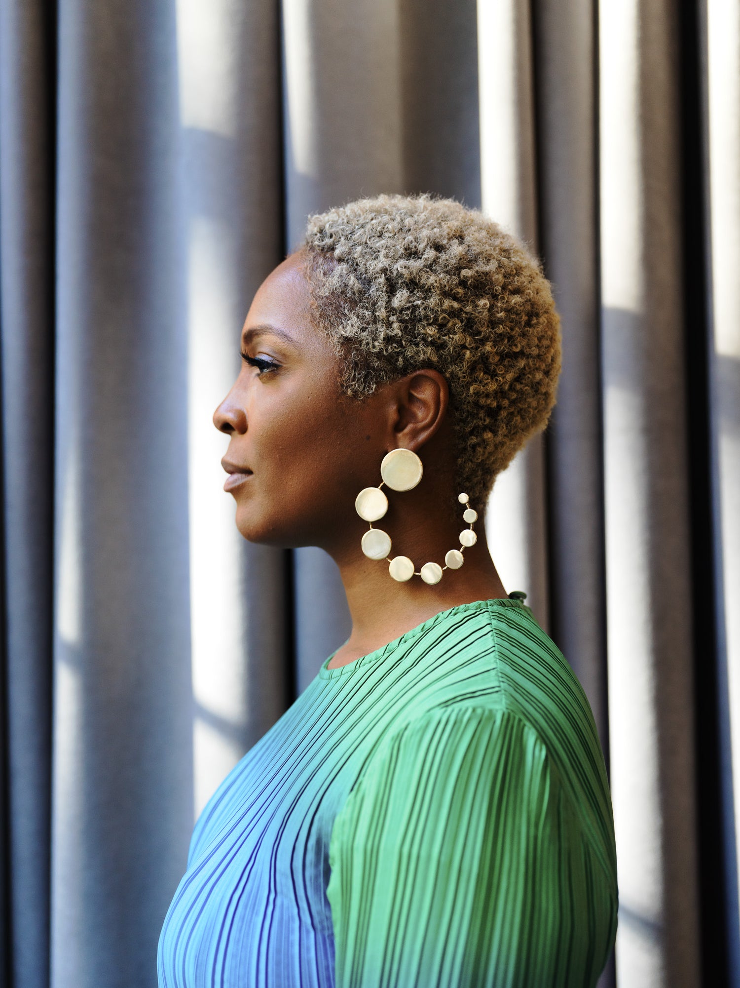 Jewelry designer Malyia McNaughton poses in profile wearing a pleated blue and green dress and bold circular statement earrings, standing against softly lit grey curtains. Her natural curls and poised expression highlight her modern elegance. Photographed by Toronto-based editorial photographer Nicholas Hyatt.