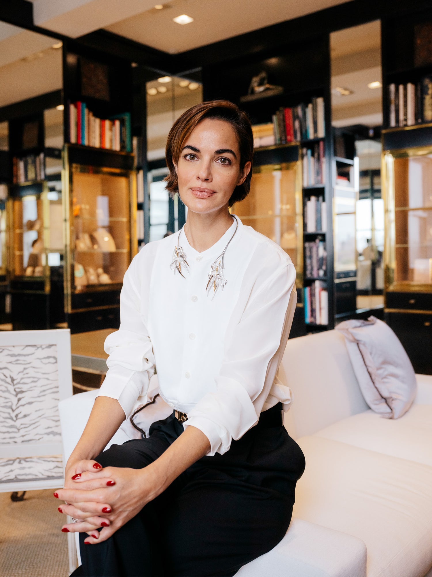 A poised Michella Cruz with short hair and natural makeup sits on a white sofa in a luxury boutique, wearing a crisp white blouse with sculptural jewelry and black trousers. Photographed by Toronto-based photographer Nicholas Hyatt, this editorial portrait captures quiet confidence and contemporary style in a refined setting.