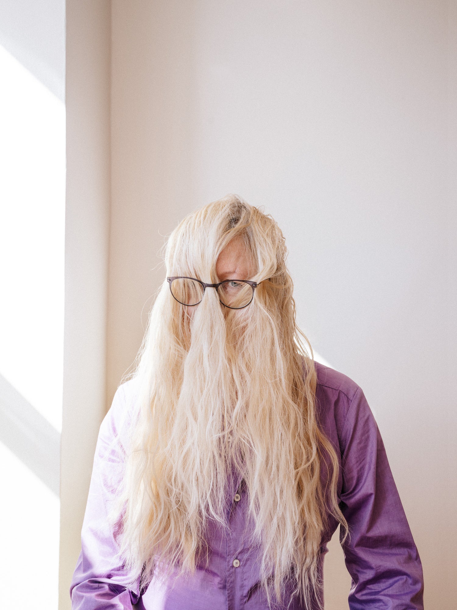 A humorous conceptual portrait Jivan Astinfalk with long platinum blonde hair draped over their face, wearing glasses and a purple button-down shirt, standing in front of a sunlit white wall. Photographed by Toronto-based photographer Nicholas Hyatt, blending wit and character-driven portraiture.