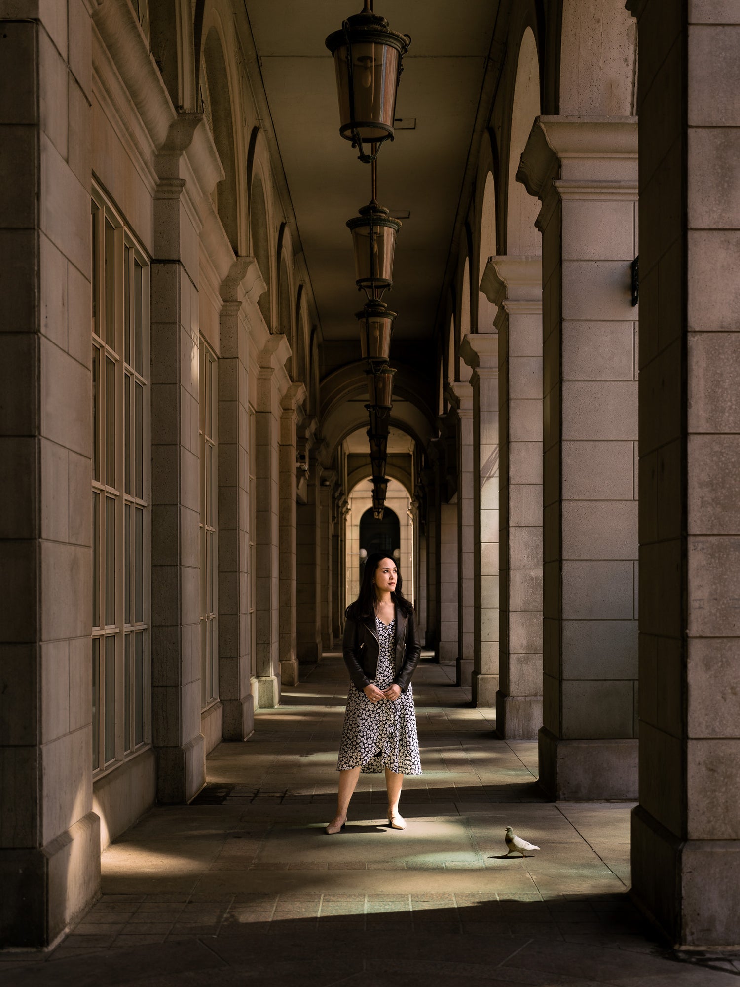  A woman in a patterned dress and leather jacket stands in a dramatic colonnade bathed in warm sunlight, her stance contemplative as a pigeon walks nearby. Photographed by Toronto-based photographer Nicholas Hyatt, this editorial portrait plays with symmetry, urban elegance, and cinematic light.